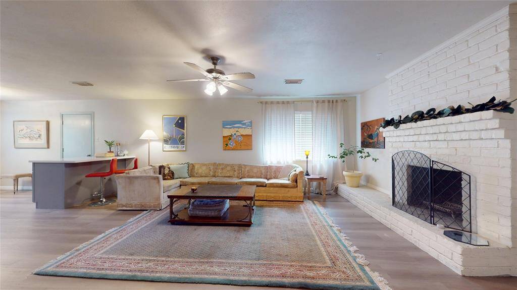 This inviting living room features a cozy seating area with a large sectional sofa and a fireplace with a white brick surround. It includes a ceiling fan, ample natural light from windows with sheer curtains, and a breakfast bar with red stools. The space is accented with artwork and houseplants, creating a warm and welcoming ambiance.