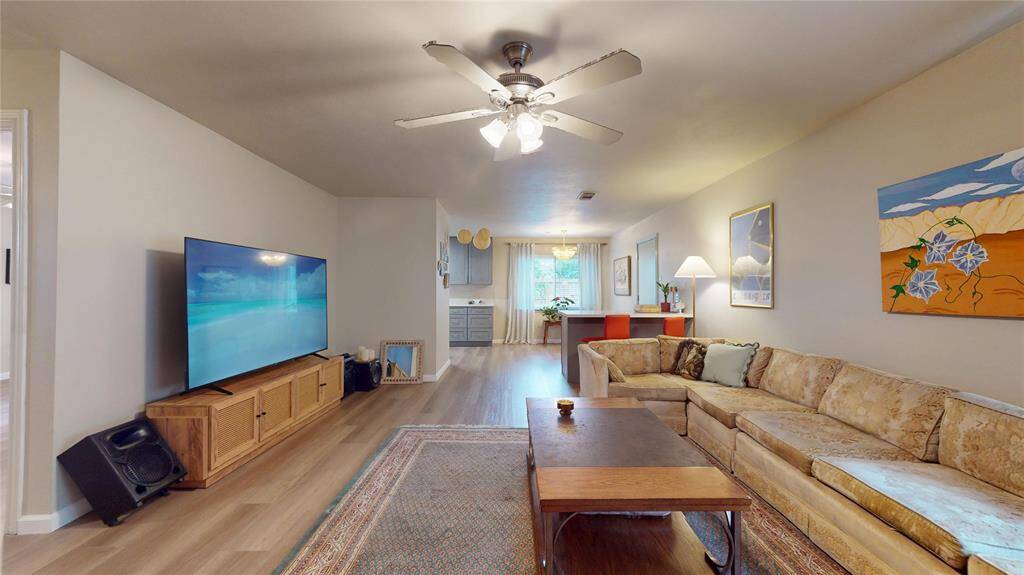 This cozy living room features a large sectional sofa, a ceiling fan, and a mounted TV. The open layout leads to a dining area with natural light, creating a welcoming space for relaxation and entertainment.