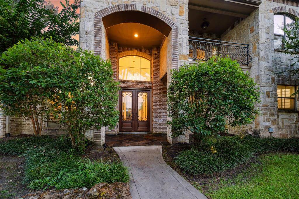 A curved walkway leads to the grand front entry, where handsome double doors with wrought iron accents set the tone for the elegant interior. The soaring stone archway and warm glow from inside create an inviting and memorable welcome.