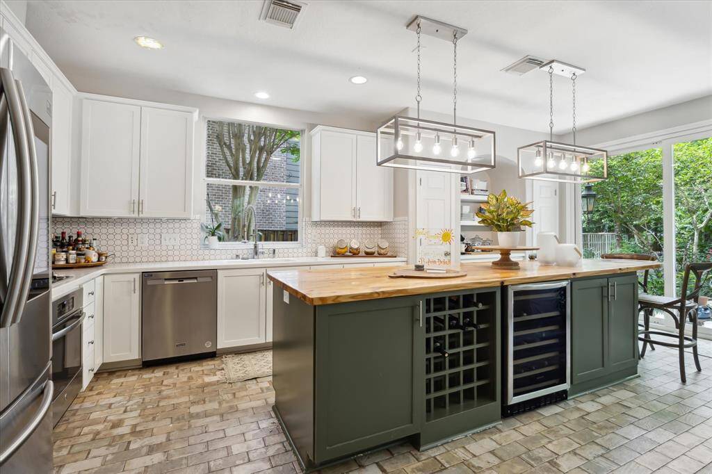 This beautifully updated kitchen blends modern style with everyday function, featuring a striking two-tone design with white upper cabinets and deep green lowers. The expanded island offers ample workspace, bar seating, a wine fridge, and built-in wine storage—ideal for entertaining. Pendant lighting adds a contemporary touch, while butcher block counters and a textured backsplash complete the warm, inviting look. Large windows and a seamless flow to the living area enhance the open-concept feel.