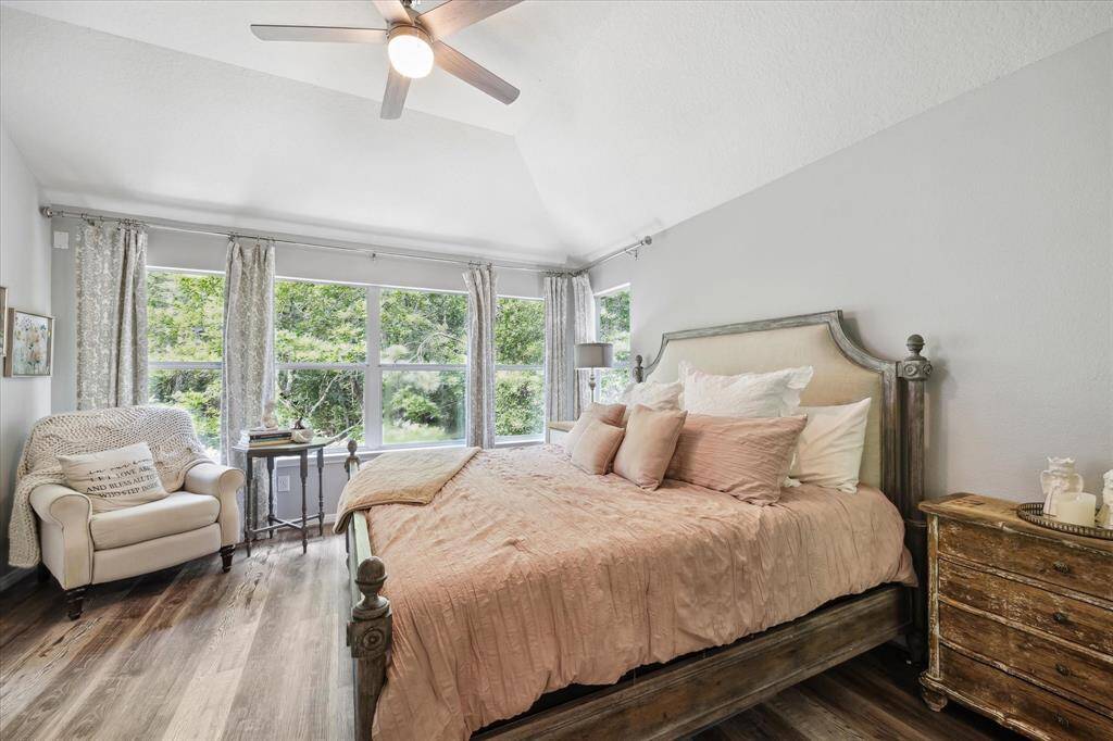 The serene primary bedroom offers a relaxing retreat with vaulted ceilings, rich vinyl plank flooring, and a wall of windows framing peaceful greenbelt views. Soft natural light fills the space, while neutral tones and elegant finishes create a calm, inviting atmosphere.
