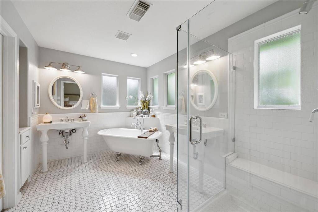 This spa-inspired primary bath blends timeless charm with modern comfort, featuring dual pedestal sinks, a freestanding clawfoot tub, and a spacious walk-in shower with built-in bench. Classic hexagon tile flooring, frosted windows for privacy, and soft natural light create a serene and elegant retreat.