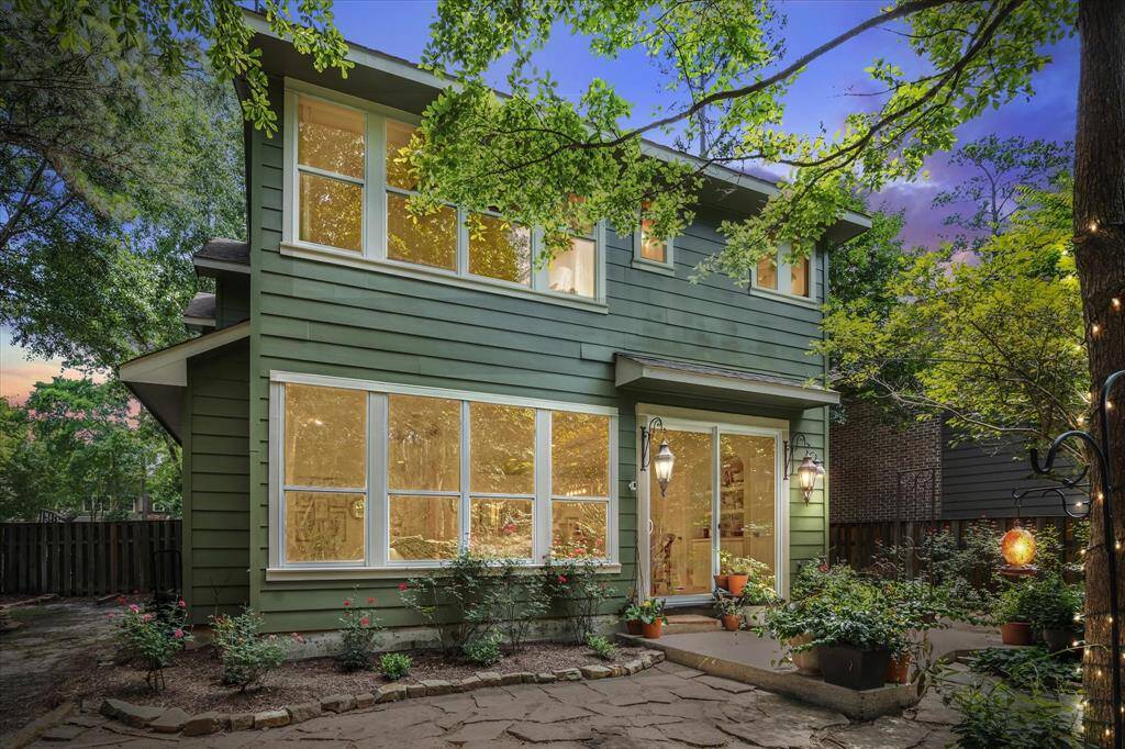 The rear elevation highlights a mix of earthy tones, with a rich green/sage exterior that blends seamlessly into the natural surroundings. Framed by mature trees and vibrant landscaping, this inviting backdrop features expansive windows and sliding glass doors that open to a charming flagstone patio—perfect for outdoor relaxation or entertaining.