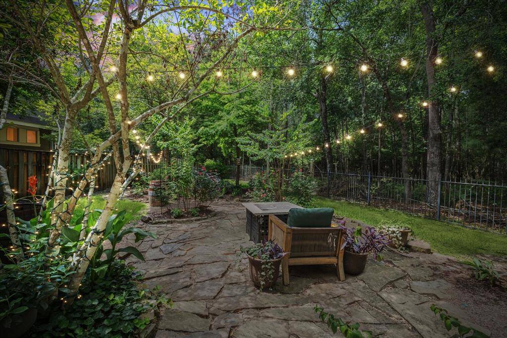 This peaceful backyard retreat is framed by a wrought-iron fence and mature trees, creating a seamless connection to the adjacent greenbelt. String lights overhead add charm and ambiance, while the flagstone patio offers the perfect setting for relaxed outdoor living—whether gathered around the fire pit or enjoying the surrounding garden beds. Lush plantings and blooming roses complete the inviting atmosphere.