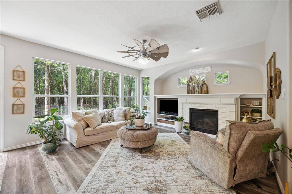 This light-filled living room offers a serene retreat with expansive windows overlooking the wooded greenbelt, filling the space with peaceful views and natural light. Rich vinyl plank flooring, a cozy gas fireplace framed by classic white millwork, and built-in shelving create warmth and function, while the unique windmill-style ceiling fan adds a stylish touch.