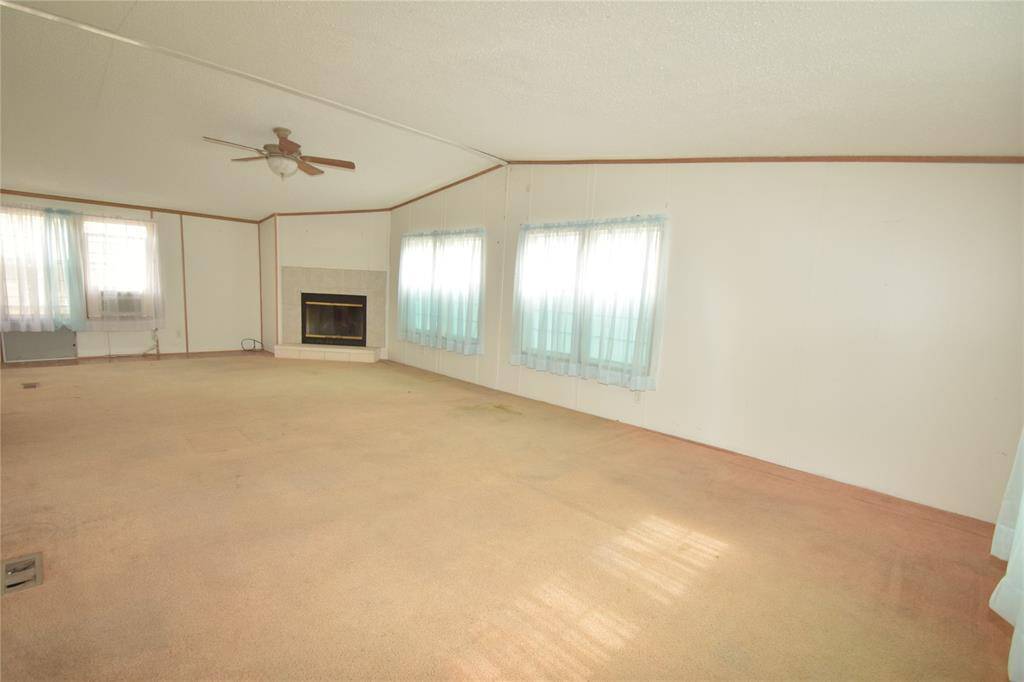 This property features a spacious manufactured home with a reddish-brown front deck, set on a large, grassy lot with mature trees. There's a covered carport on the side, providing convenient vehicle storage.