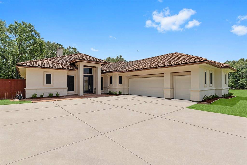 This modern home features a spacious driveway leading to a three-car garage. It has a Mediterranean-style design with a red-tiled roof, light-colored exterior walls, and large windows. The property is surrounded by lush greenery, offering privacy and a serene atmosphere.