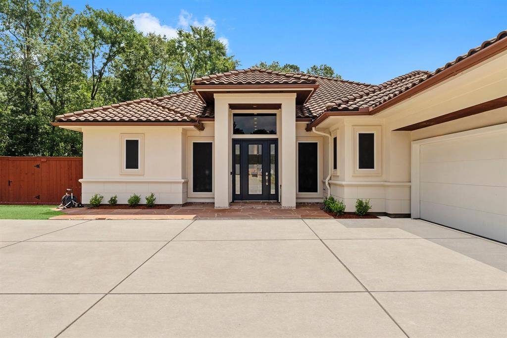This photo showcases a modern, single-story home with a Mediterranean style, featuring a clean, spacious driveway, elegant tile roofing, and a welcoming entrance with large windows. The property is enhanced by a well-maintained yard and surrounded by lush greenery, providing a serene and inviting atmosphere.