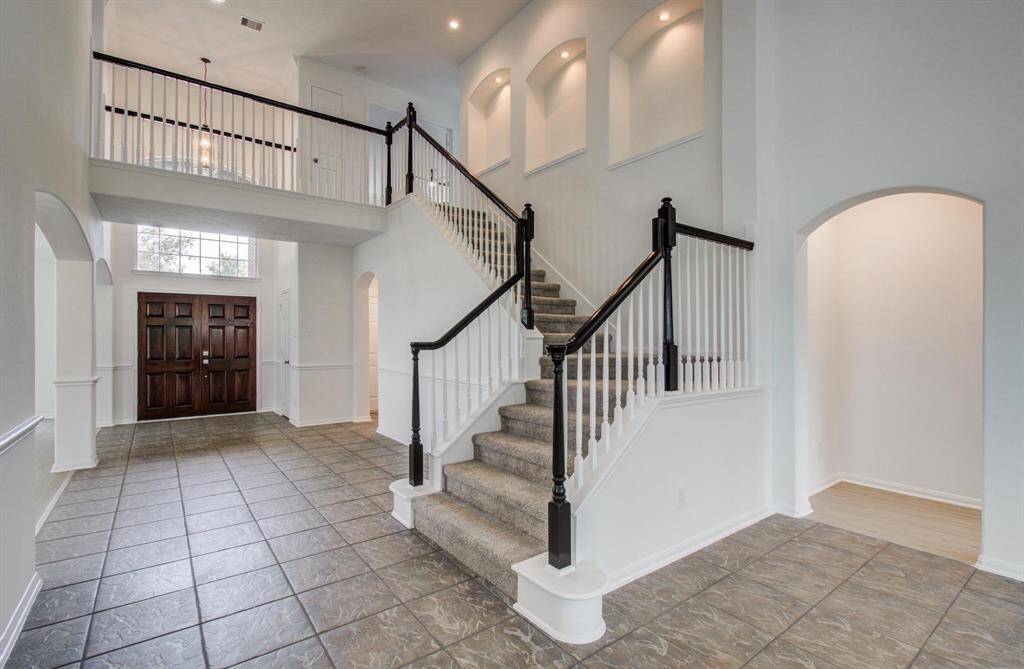This elegant foyer features a grand staircase with dark wood accents and white railings, high ceilings, and ample natural light from large windows. The tiled floor and spacious layout create a welcoming entrance.