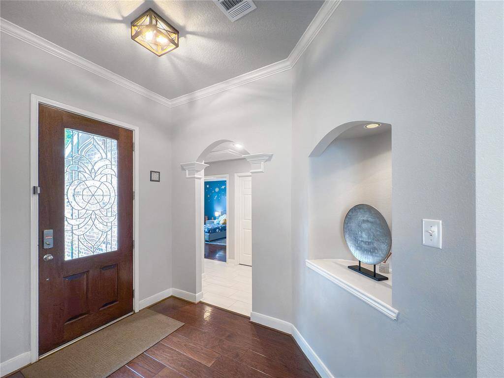 This entryway features a rich wooden door with intricate glass detailing, leading into a welcoming foyer with hardwood floors. Elegant archways and a decorative niche add charm, while a modern ceiling light enhances the space.