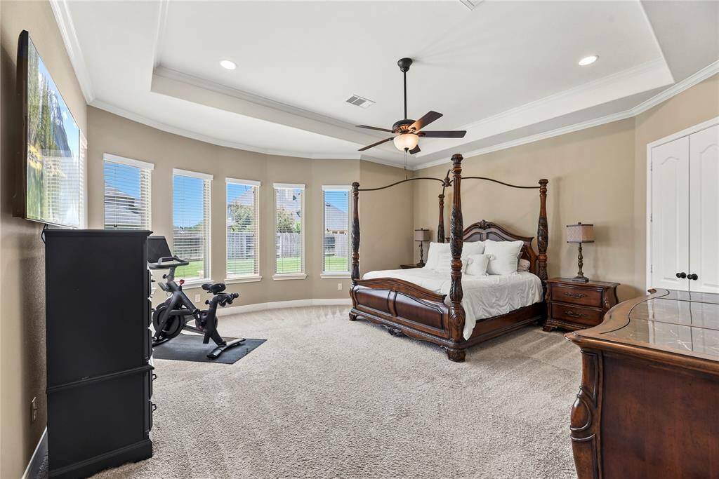 Spacious first-floor primary bedroom measuring 18.11 x 18.3, featuring a tray ceiling, multiple windows with serene backyard views, and cozy carpet flooring.