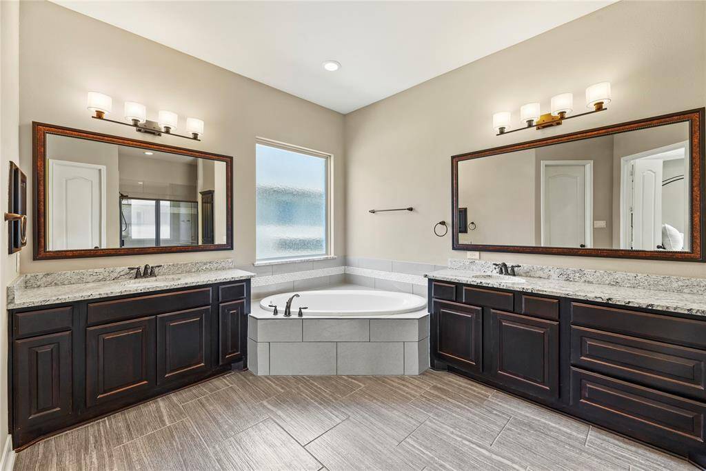 Spacious primary bathroom featuring dual vanities with granite countertops, large mirrors, and double sinks. It includes a corner soaking tub, tile flooring, and a frosted window for privacy.