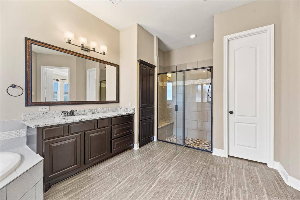 This spacious primary bathroom features a sleek design with a large vanity, a granite countertop, and it includes a glass-enclosed shower.
