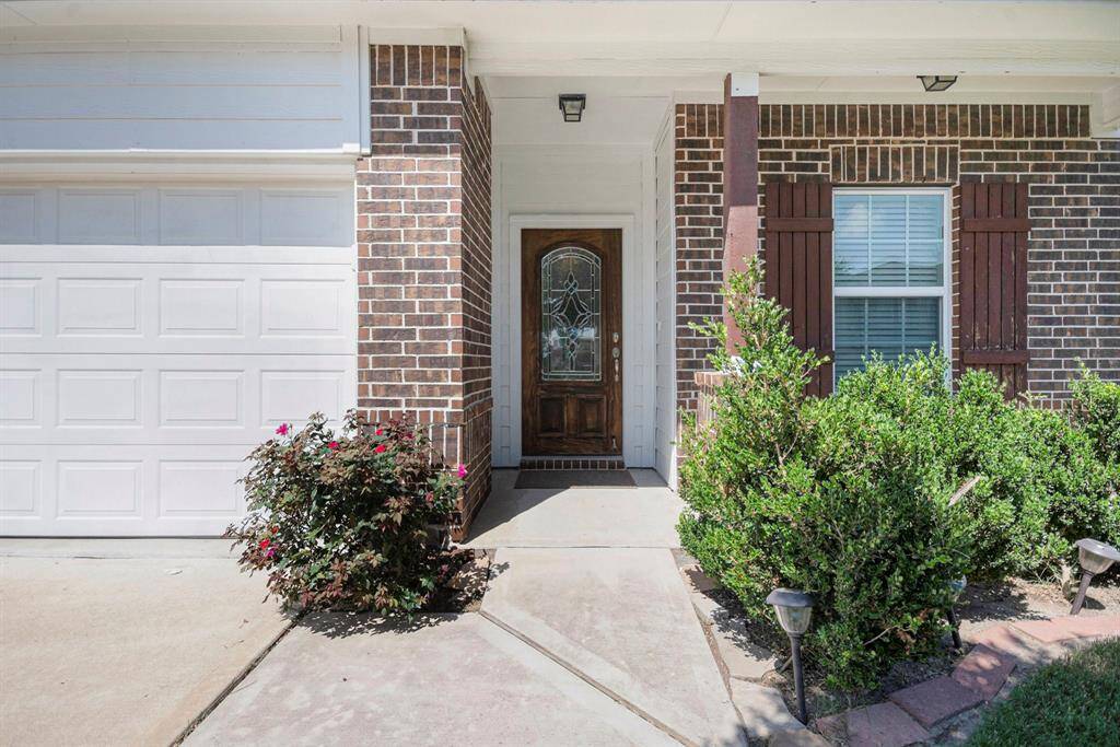 Welcome to your charming entryway, featuring a stylish brick facade and elegant glass door, framed by lush greenery.