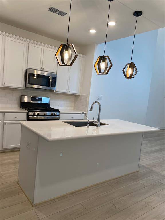 This photo showcases a spacious, modern kitchen and living area with high ceilings and large windows. The kitchen features white cabinetry, a sleek island with a sink, and stainless steel appliances. Light wood flooring and ample natural light create an open, airy feel.