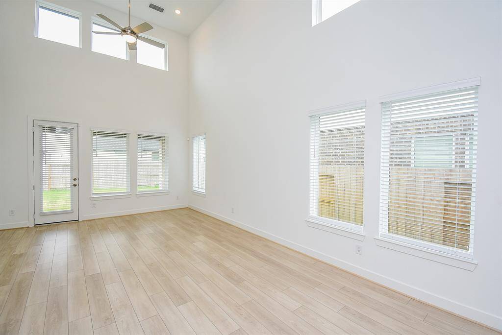 This spacious, bright room features high ceilings, large windows with blinds, a ceiling fan, and light wood flooring. There's a door leading to the backyard, enhancing indoor-outdoor living.