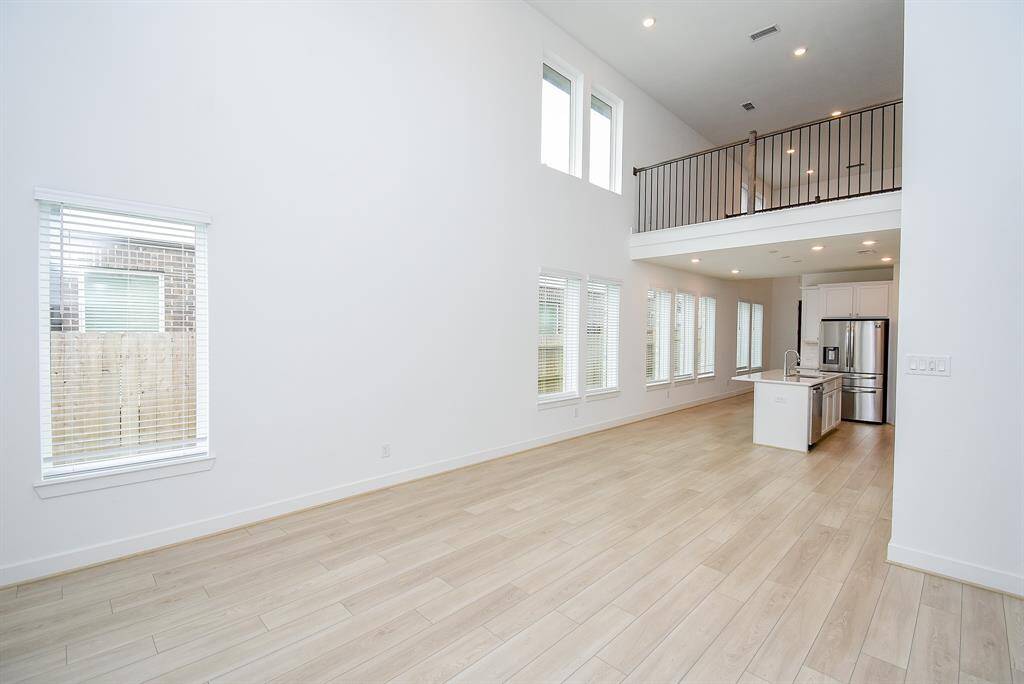 This photo showcases a bright, spacious open-concept living area with high ceilings, large windows, and light wood flooring. The kitchen features modern appliances and an island, while a loft area overlooks the space, adding an airy, contemporary feel.