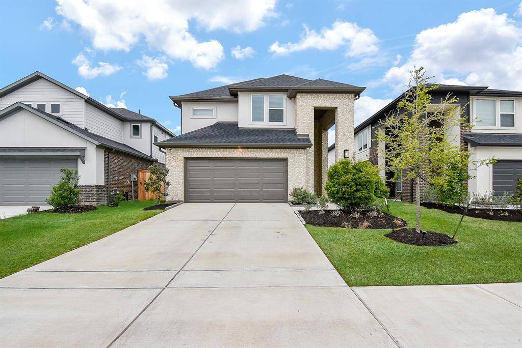 This photo showcases a modern two-story home with a brick and siding exterior. It features a spacious driveway leading to a double garage, and well-maintained landscaping with small trees and shrubs. The neighborhood appears to be newer and well-kept.