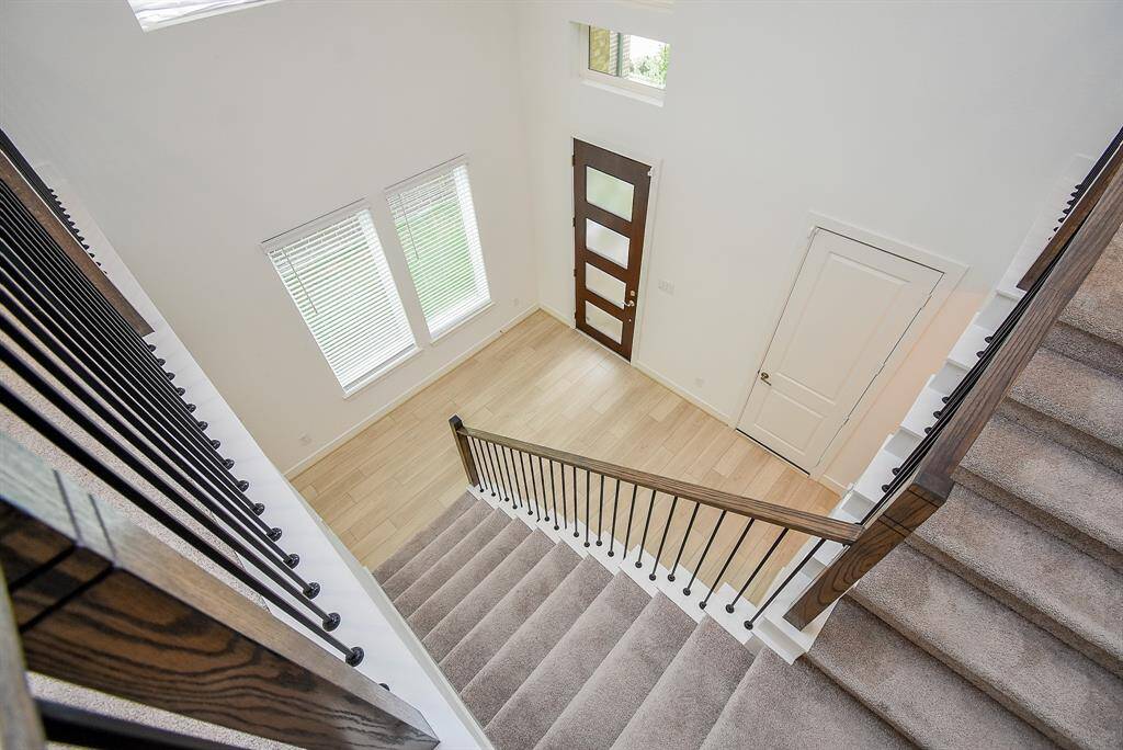 This photo shows a bright, open entryway with high ceilings. The staircase features carpeted steps and a sleek wooden railing with metal spindles. Natural light floods in through large windows and the modern front door with frosted glass panels, complementing the light wood flooring.