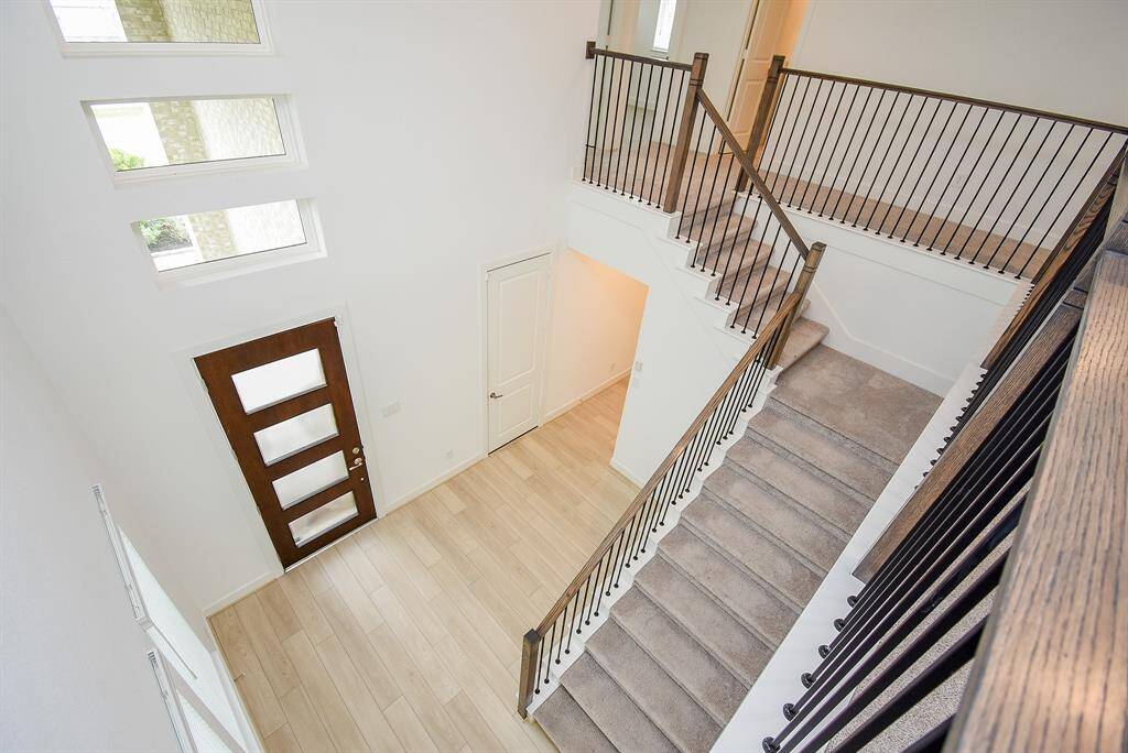 This photo showcases a modern, bright entryway with a stylish wooden door featuring glass panels. The space includes a staircase with carpeted steps and sleek metal railings, opening up to a second-floor landing. The flooring is light, enhancing the airy feel.