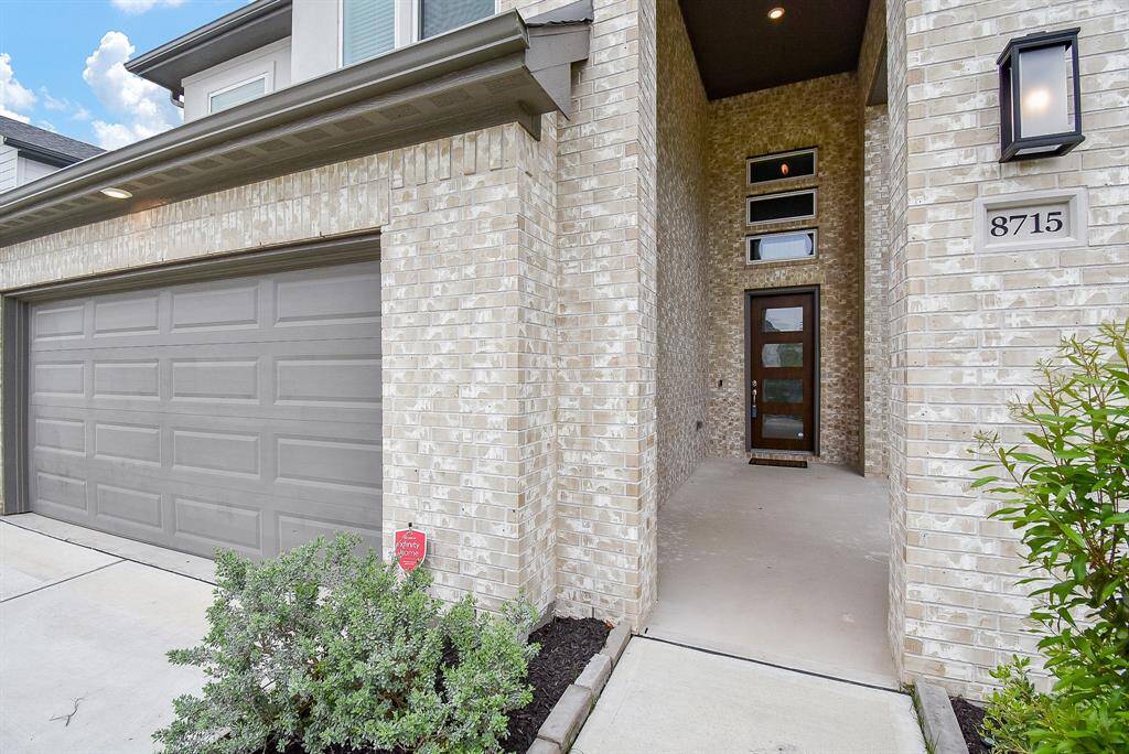 This photo showcases a modern brick home with a two-car garage and a welcoming recessed entryway. The clean, contemporary design includes a stylish door with glass panels, surrounded by well-maintained landscaping.