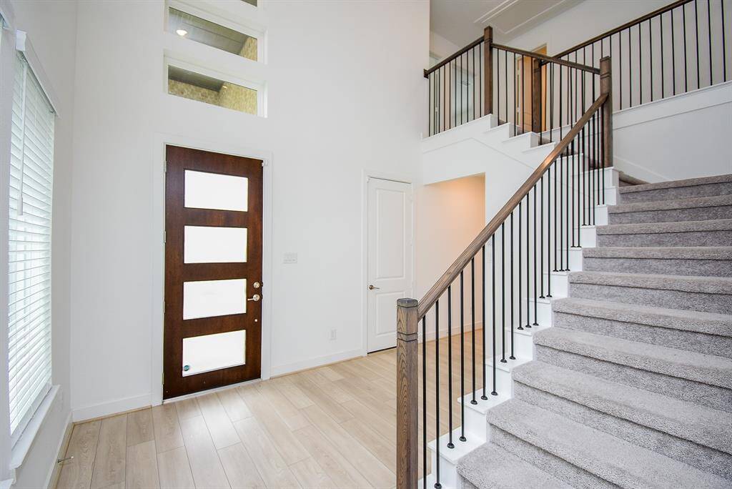 This photo showcases a bright, modern entryway with a dark wood door featuring frosted glass panels, high ceilings, and a carpeted staircase with sleek black railings, leading to an upper level. Light wood flooring and ample natural light enhance the space's openness.