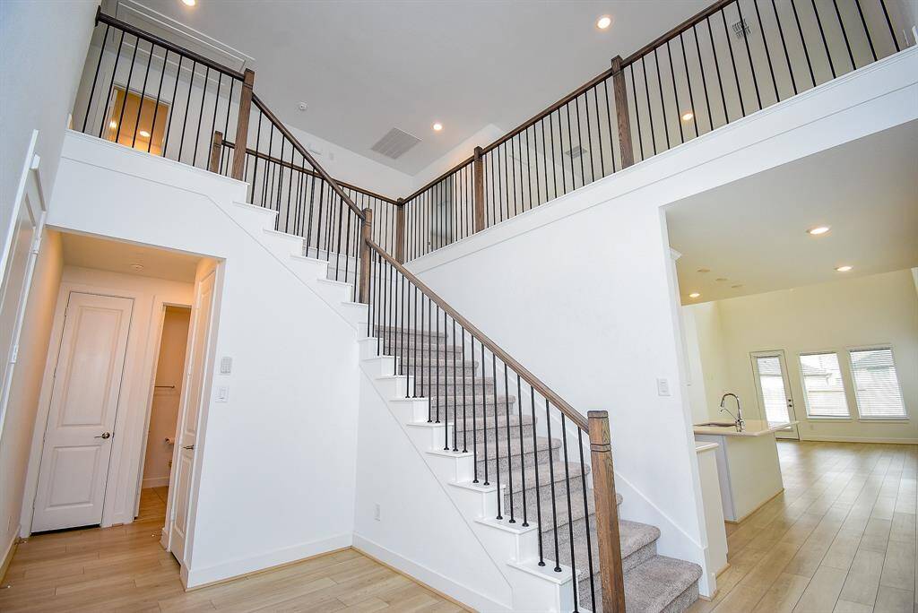 This photo showcases a bright, modern interior with an open staircase featuring dark railings, leading to an upper level. The space has light wood flooring, recessed lighting, and a kitchen area with a sleek design. Large windows provide ample natural light, creating an airy and welcoming atmosphere.
