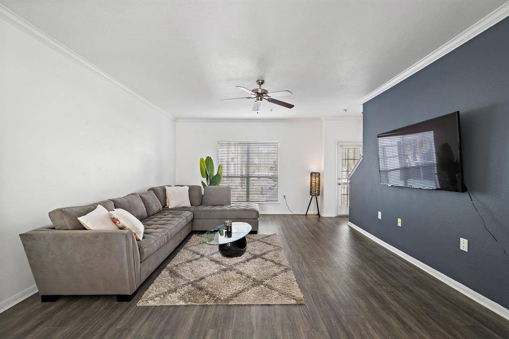 Spacious living room with dark wood flooring and spacious enough to accomendate a large sectional sofa or multiple sitting areas. It has a ceiling fan, and natural light from a window and glass door, offering a cozy yet contemporary feel.