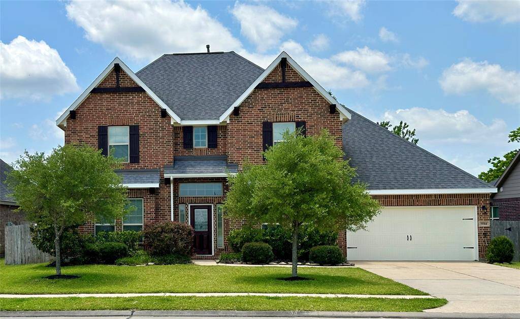 Charming two-story brick home with a gabled roof, featuring a well-maintained front yard with lush greenery and two trees. The house includes a spacious driveway leading to a 3 car garage.