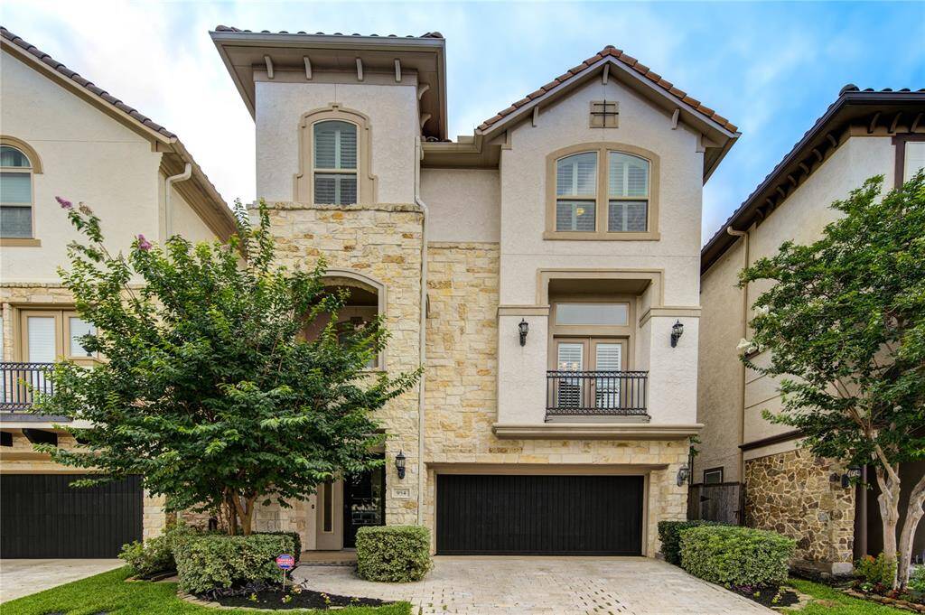 This is a modern, three-story home with a stone and stucco exterior, featuring a two-car garage and small landscaped front yard. It has arched windows, decorative wrought iron accents, and is nestled between similar style homes.