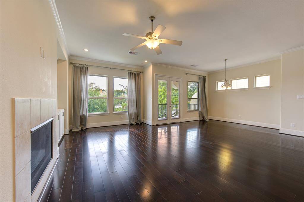 Spacious living room with dark hardwood floors, large windows, and a ceiling fan. Features a fireplace, ample natural light, and a door leading to a balcony, offering a bright and inviting atmosphere.