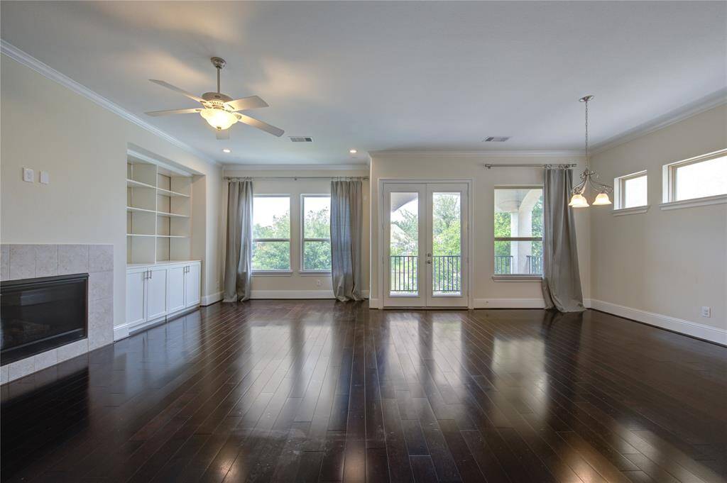 Spacious living area with dark hardwood floors, a cozy fireplace, built-in shelving, and large windows offering ample natural light. Features recessed lighting and an open layout with a view of the staircase.