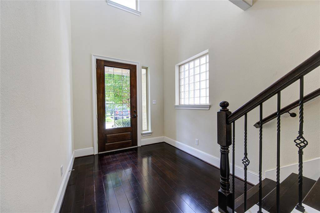 Walk right into a bright entryway with dark hardwood floors, a wooden front door with glass panels, and a staircase with wrought iron railings. The space is well-lit with natural light from a high window and a glass block window.