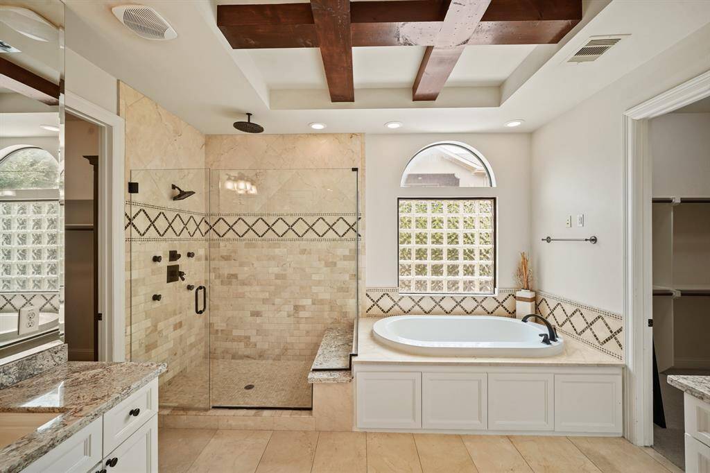 This spa-like bath features a tile surrounded whirlpool tub with arched privacy glass window above and nearby over-sized walk-in frameless glass shower with multiple wall & ceiling mounted shower heads surrounded by travertine tile.