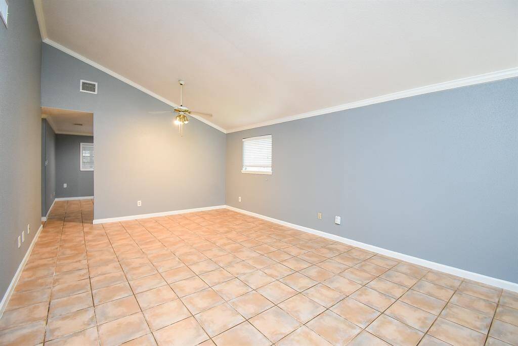 This spacious room features a high ceiling with a ceiling fan, light gray walls, and large tile flooring. Natural light enters through a window, creating a bright and airy atmosphere.