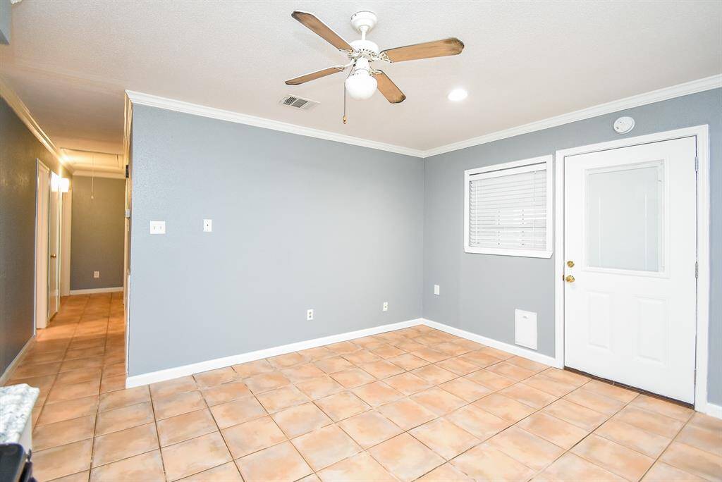 Bright room with light gray walls and terracotta floor tiles. It features a ceiling fan, white trim, and a small window next to a door leading outside. The space connects to a hallway.