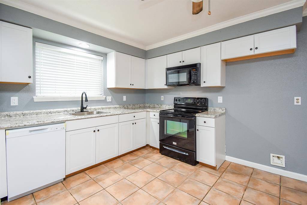 This kitchen features modern white cabinetry with granite countertops, a black stove and microwave, and a dishwasher. It has a clean, bright look with tiled flooring and a large window for natural light.