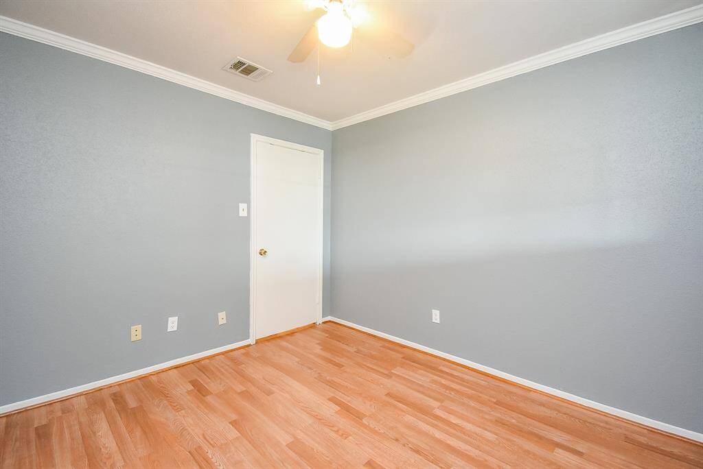 This photo shows a bright, empty room with light wood flooring and soft gray walls. It features a ceiling fan and crown molding for added detail. The room has a single door and electrical outlets along the baseboards, offering a clean slate for personalization.