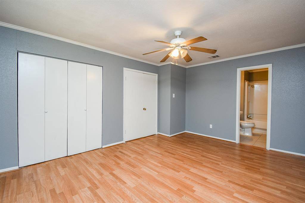 This spacious room features laminate wood flooring, soft gray walls, and a ceiling fan. It includes a closet with white sliding doors and an en-suite bathroom with a bathtub.