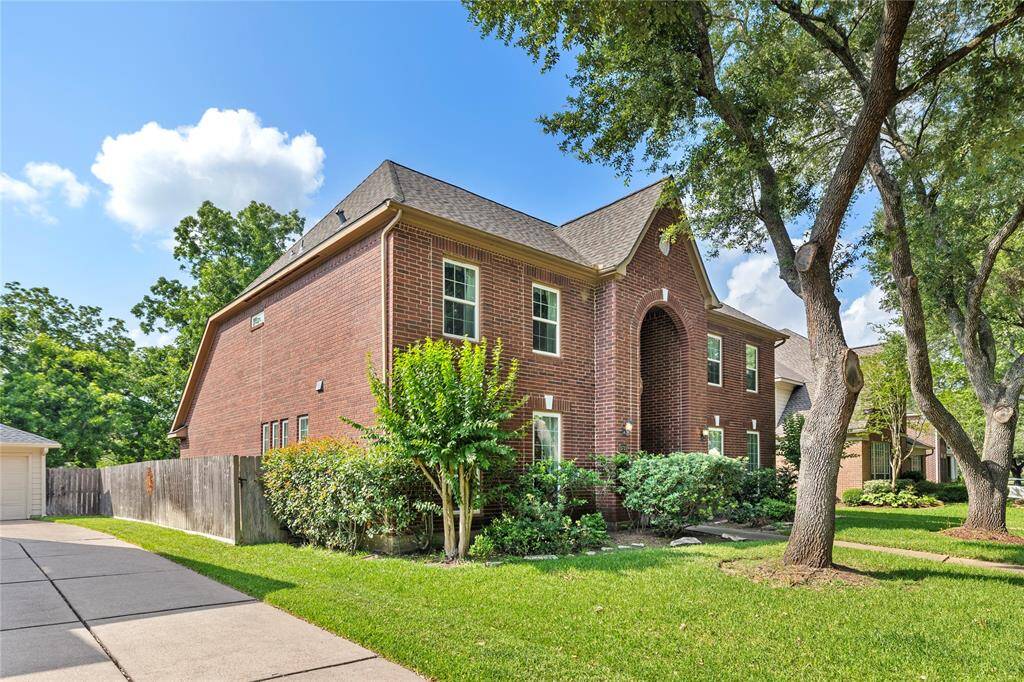 This photo shows a charming two-story brick home with a well-maintained lawn and mature trees. The property features a spacious driveway and is surrounded by a wooden fence, offering privacy and a welcoming atmosphere.