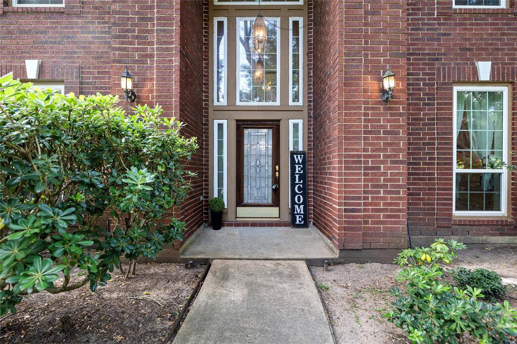 Welcoming entryway with a brick facade, large windows, and elegant lighting. A clear path is flanked by greenery, leading to a decorative glass front door.
