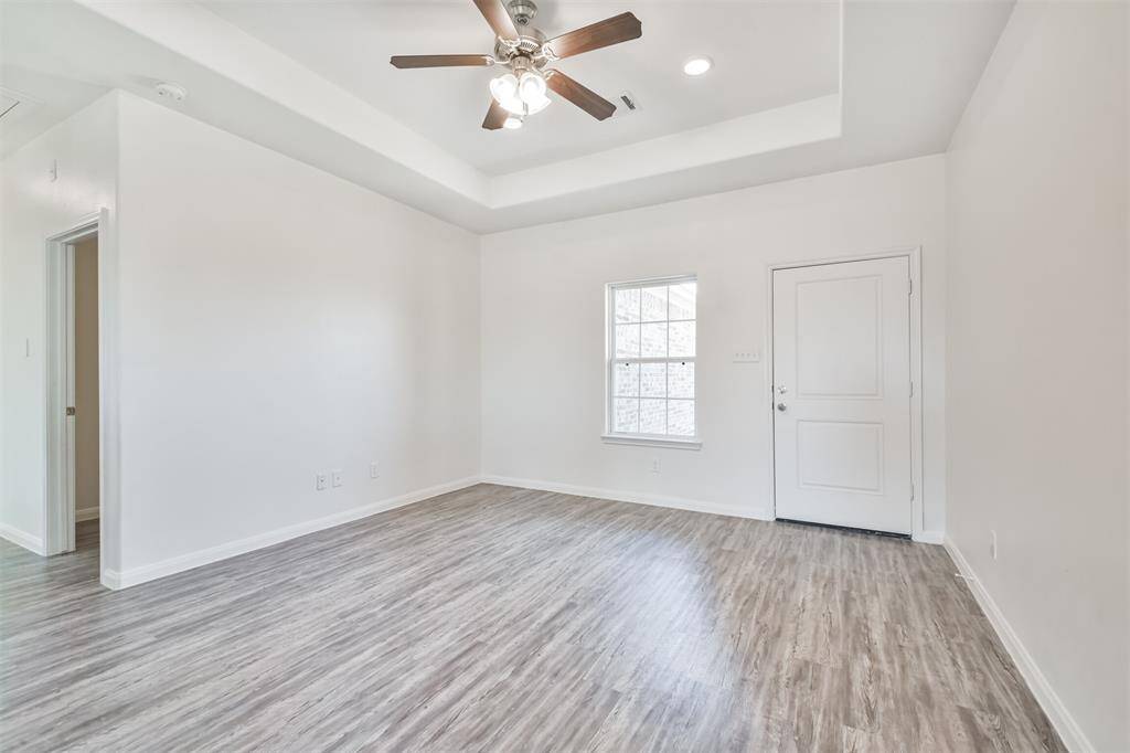 This room features a modern design with light wood-style flooring, a ceiling fan with lighting, and a recessed ceiling. It has a window for natural light and a door leading to another area, offering an inviting space for customization.