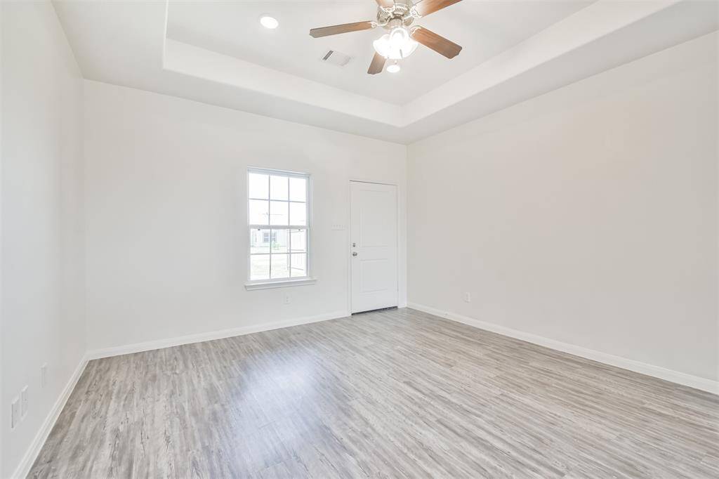 Bright, spacious room with light wood flooring, tray ceiling, ceiling fan, and large window providing natural light.