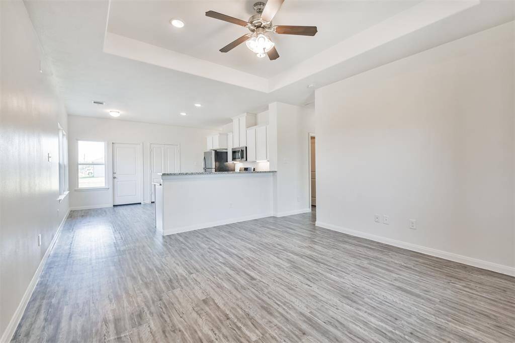 This photo showcases a bright, open-concept living area with a modern kitchen. It features light wood flooring, white walls, and a ceiling fan. The kitchen has stainless steel appliances, white cabinetry, and a breakfast bar. Natural light enters through a window, enhancing the airy feel.