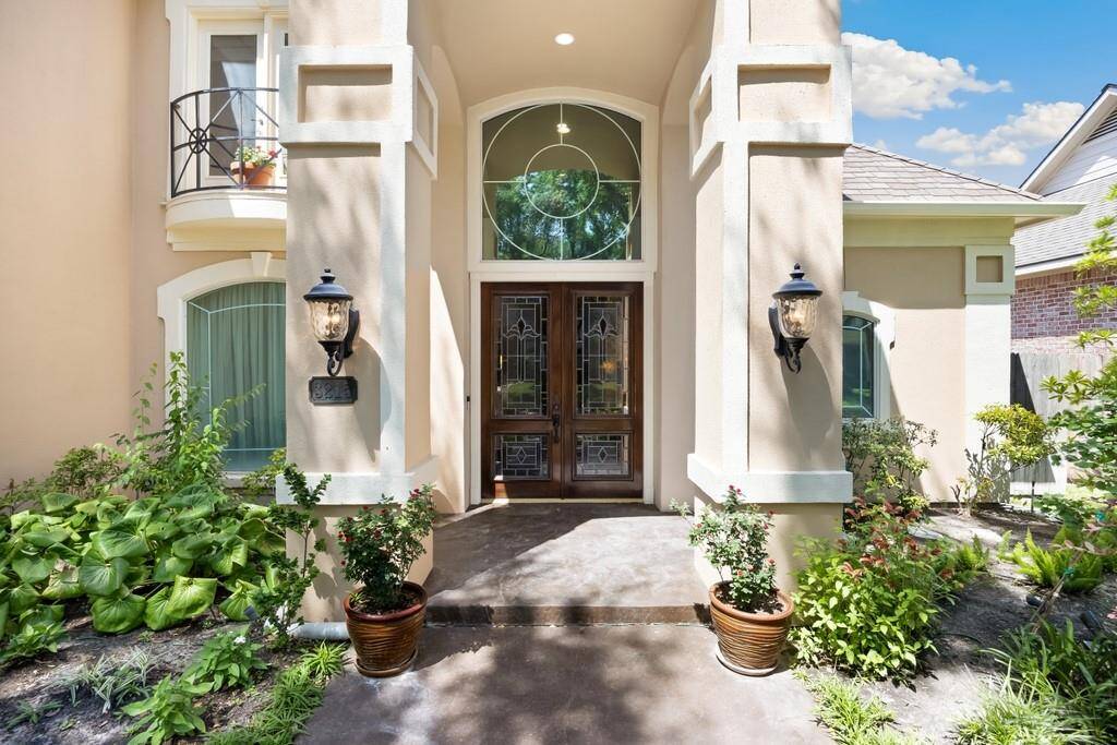 A sophisticated entryway welcomes guests with stately columns, architectural accents, and double glass doors framed by lush gardens. The grand arched transom window above sets the tone for the elegant interiors within.