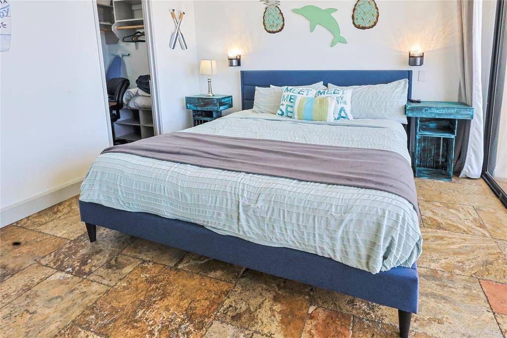 The bedroom features a beach-themed decor with warm walls and ocean-inspired accents. It includes a comfortable bed with blue and white bedding, turquoise nightstands, and wall art. Natural light fills the room, creating a bright and inviting atmosphere. The door leads to the large walk in closet.