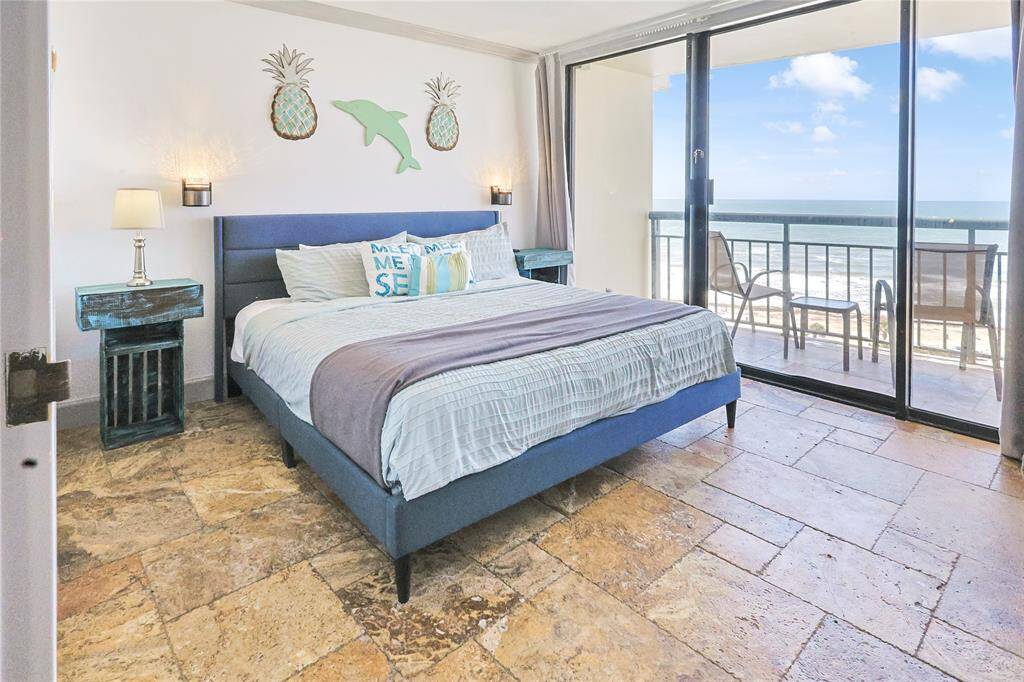 Another view of the bright bedroom featuring a king sized bed with cushioned pillows and stylish blue side tables. It opens up to a balcony with ocean views through sliding glass doors, offering abundant natural light.