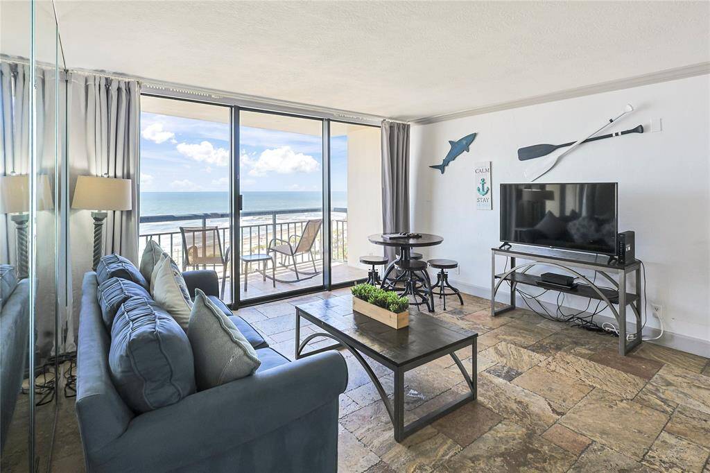 This beautiful living room features a bright, open layout with a large sliding glass door leading to a balcony with Gulf views. It’s furnished with a plush blue sofa, a wooden coffee table, and a TV setup. A beautiful dining area with high stools adds functionality, while beach-themed decor creates a relaxed, inviting atmosphere.