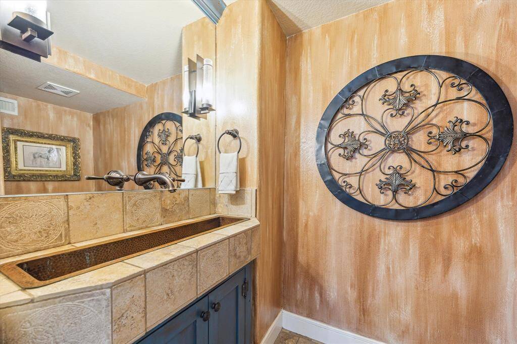 Frist floor powder room with stone counter and inset metal trough sink, wall faucet and sconces inset into mirror, and decorative painted wall and cabinet.