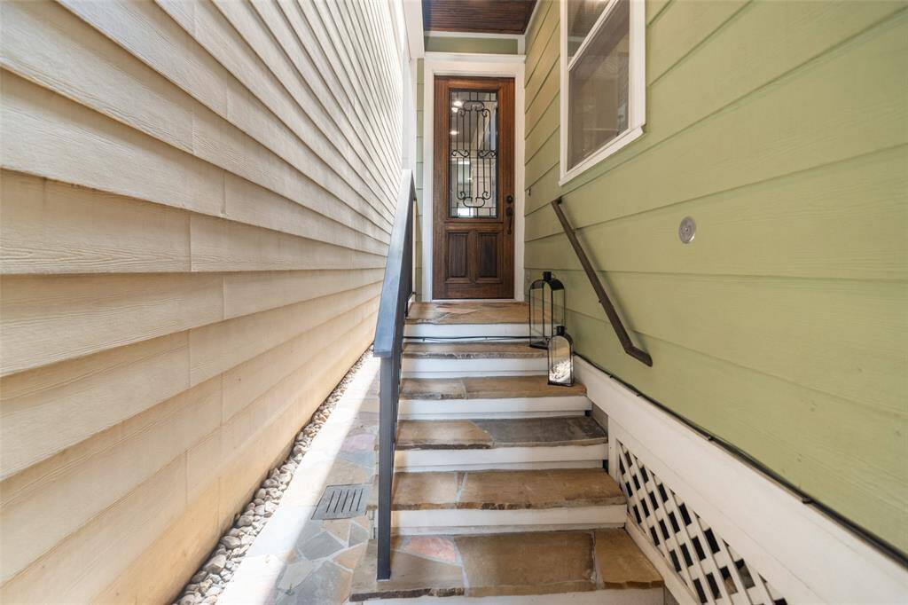 This photo shows a narrow entryway with steps leading to a wooden front door. The path is bordered by beige siding on one side and green siding on the other, with a decorative lantern placed beside the stairs.